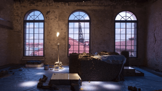 A dimly lit room with big windows, a candle lit, plastered walls, and a large block of concrete